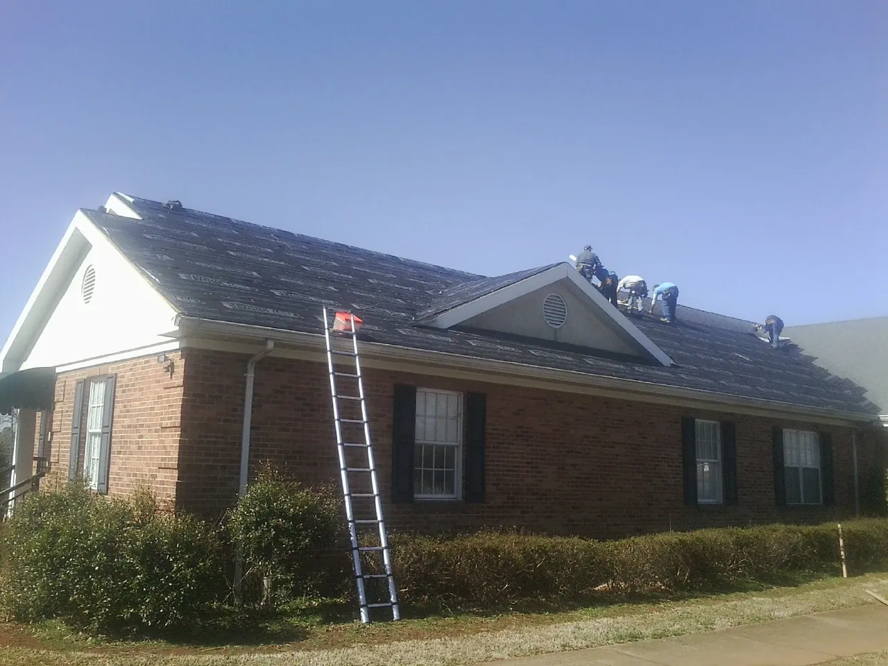 Crew tearing shingles off a brick ranch roof with a ladder set — Emergency Roof Repair work by our crew in Irmo
