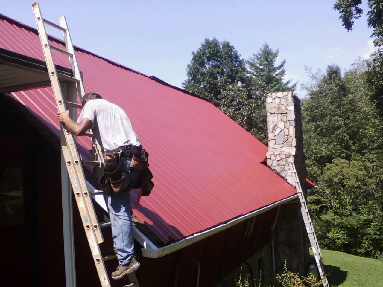 Roofer on a ladder beside a red metal roof and stone chimney — Soffit & Fascia Repair by Roofing Brothers in Irmo